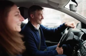 Two people are seated in the front seats of a car, with the man driving and the woman in the passenger seat smiling.